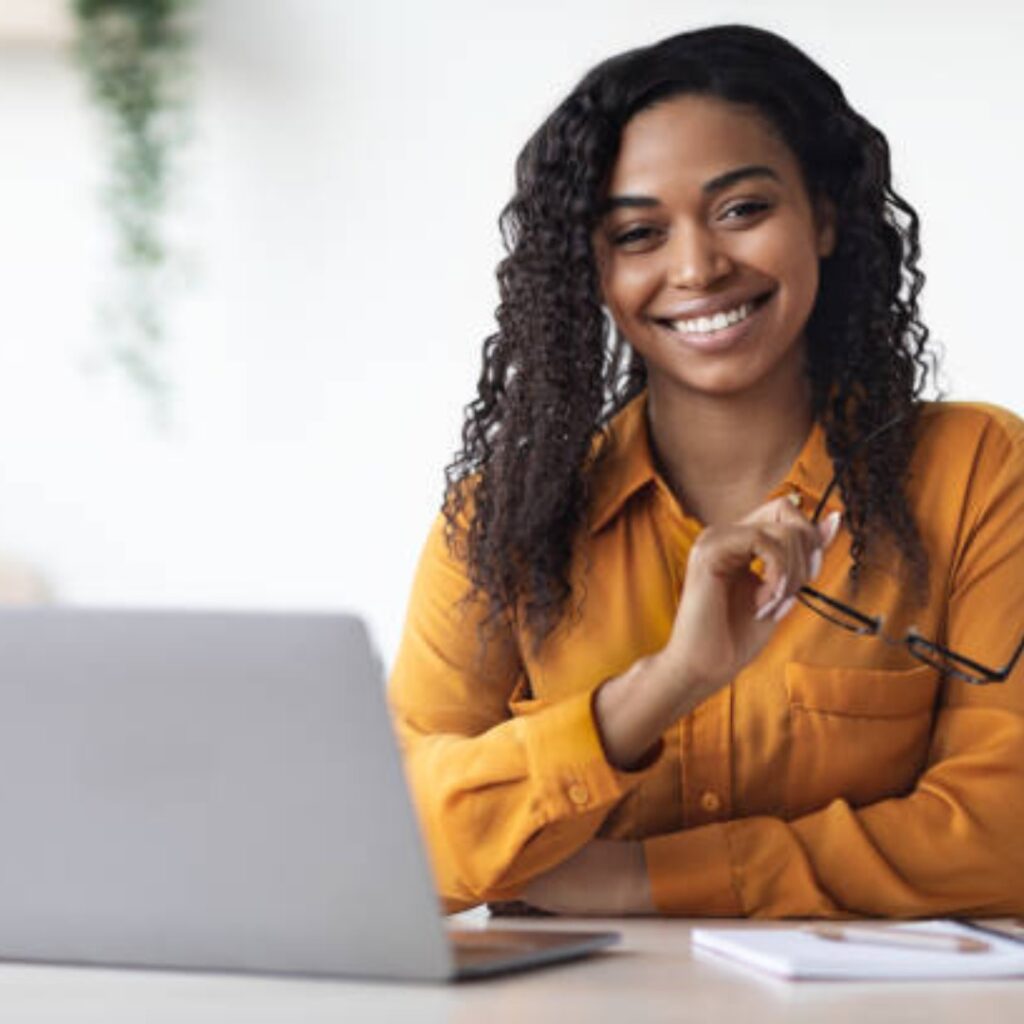 zvvt ict program promo image - smiling black woman with long curls, sitteng behind a laptop, wearing a yellow top.